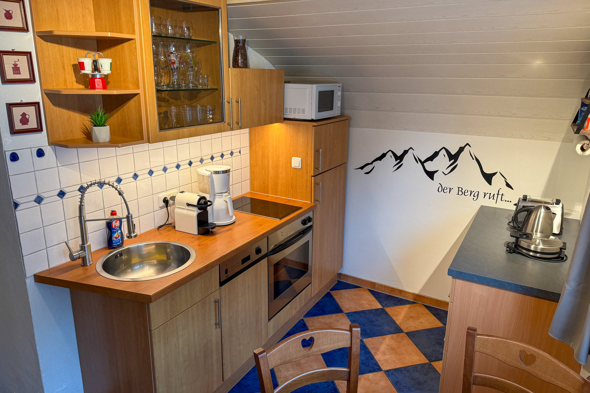 Voll ausgestattete Küche mit Holzmöbeln, Wandtattoo 'Der Berg ruft' und Kaffeemaschinen.