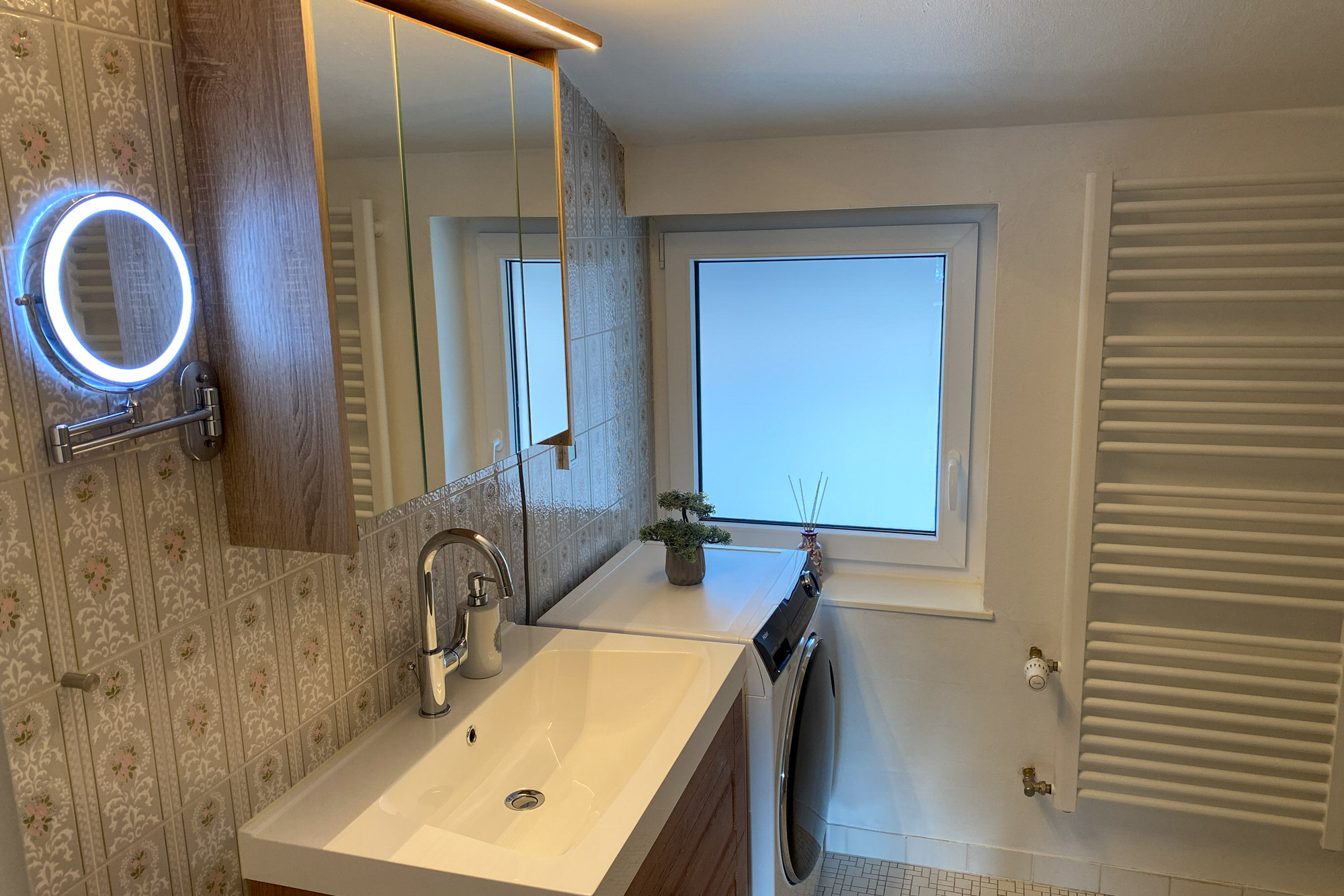 Badezimmer mit Spiegel, Waschtisch, Kosmetikspiegel und Waschmaschine.